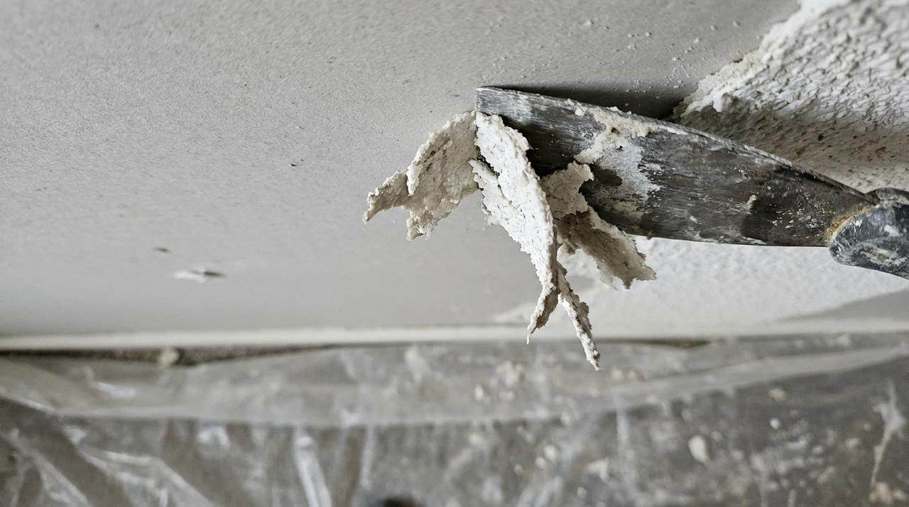 Wide drywall scraper blade lifting damp ceiling texture from drywall in a plastic-sheeted room